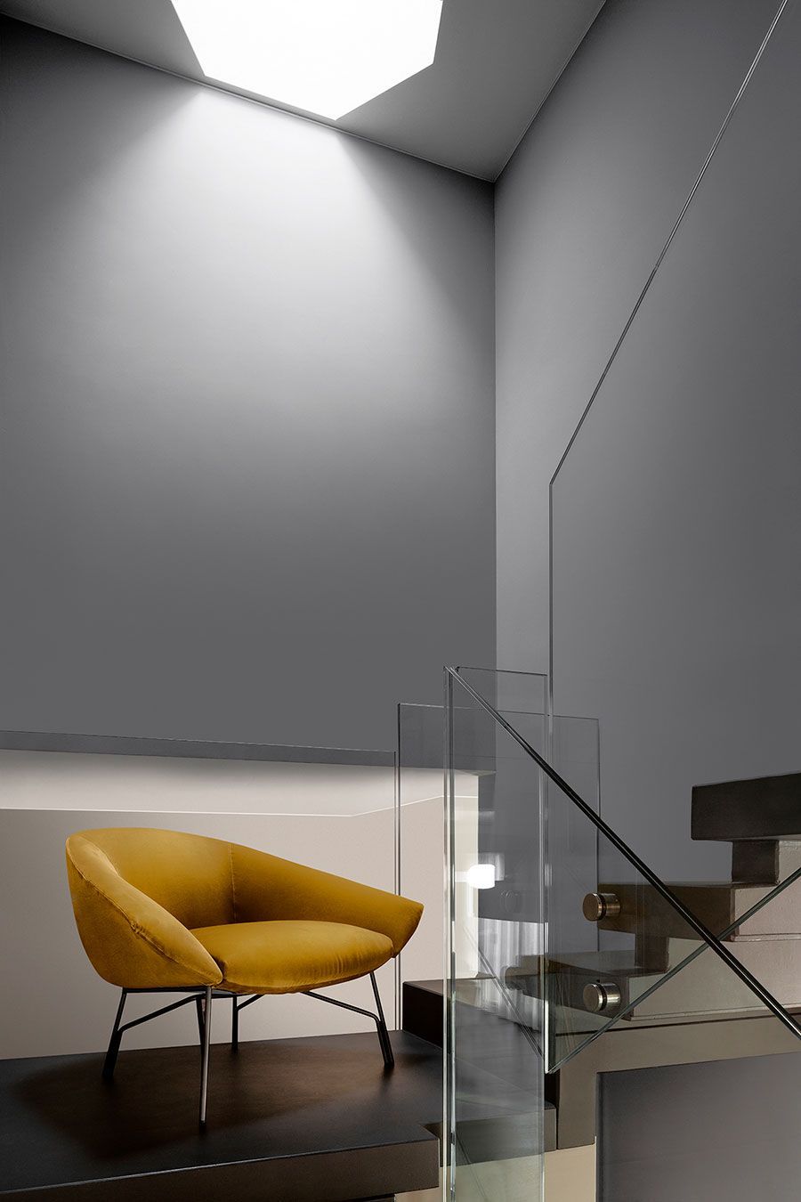 Sala de estar moderna con silla de color amarillo mostaza, escaleras de cristal y paredes altas grises.