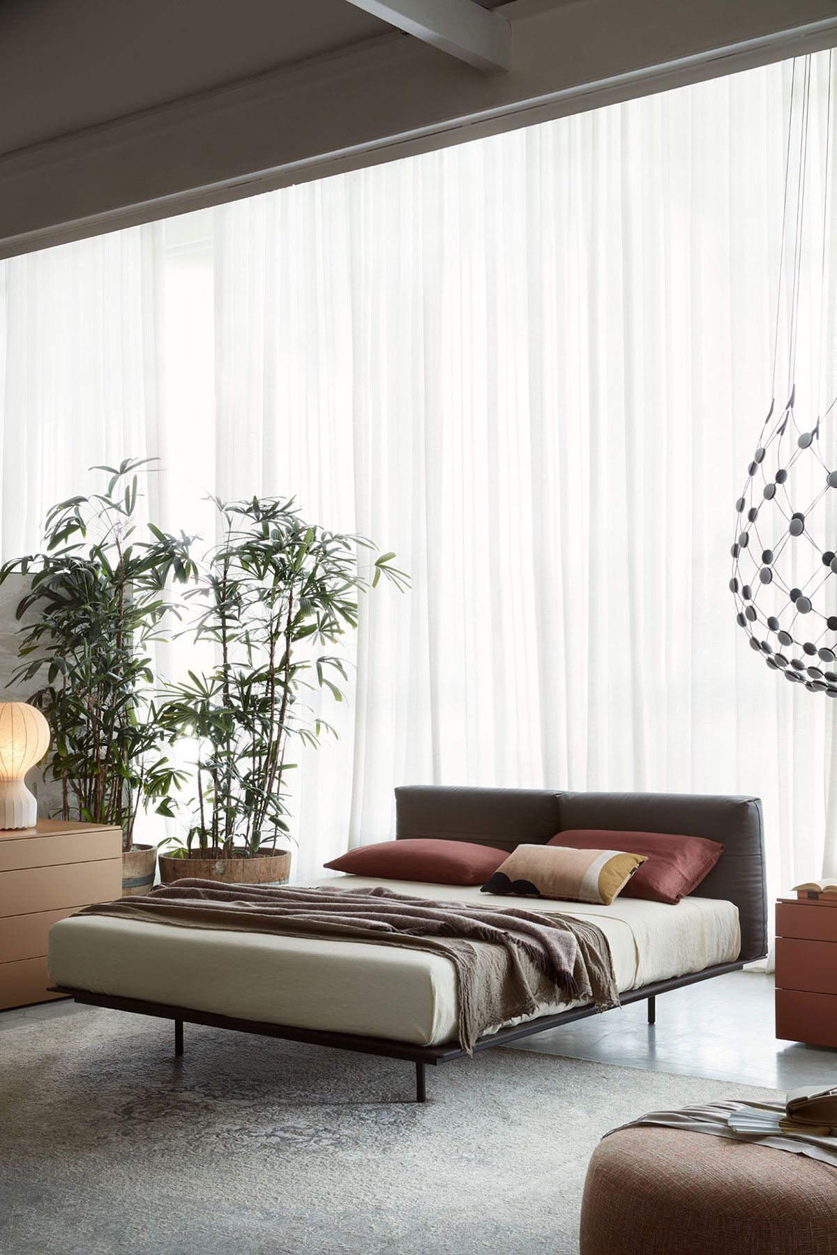 Dormitorio moderno con cama baja, plantas, cortinas transparentes y luz natural suave.