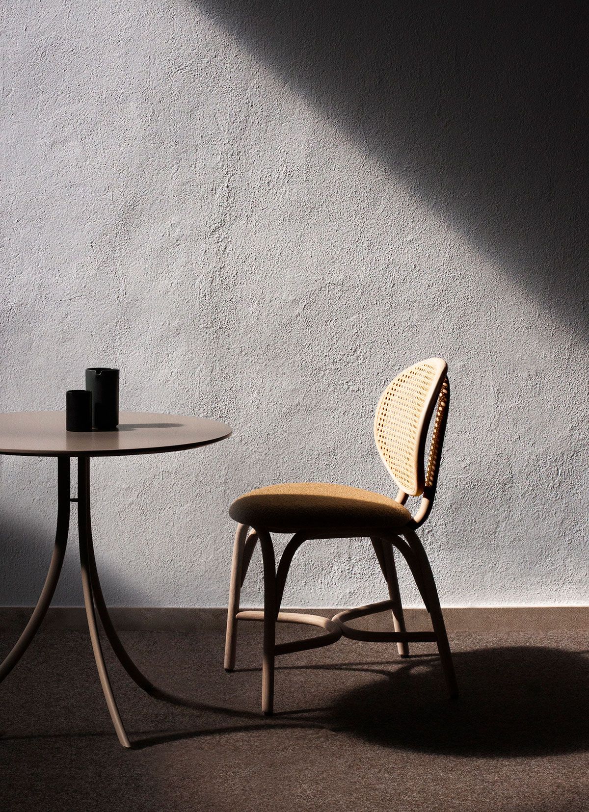 Habitación minimalista con una mesa redonda, tazas negras y una silla de madera, bañada por la luz del sol.
