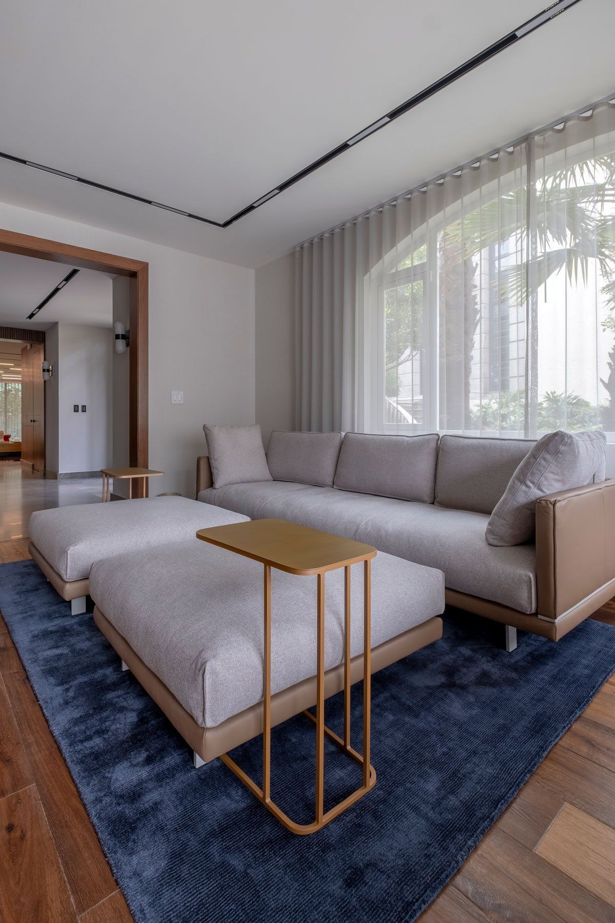 Sala de estar moderna con sofá gris, alfombra azul y grandes ventanales con cortinas transparentes.