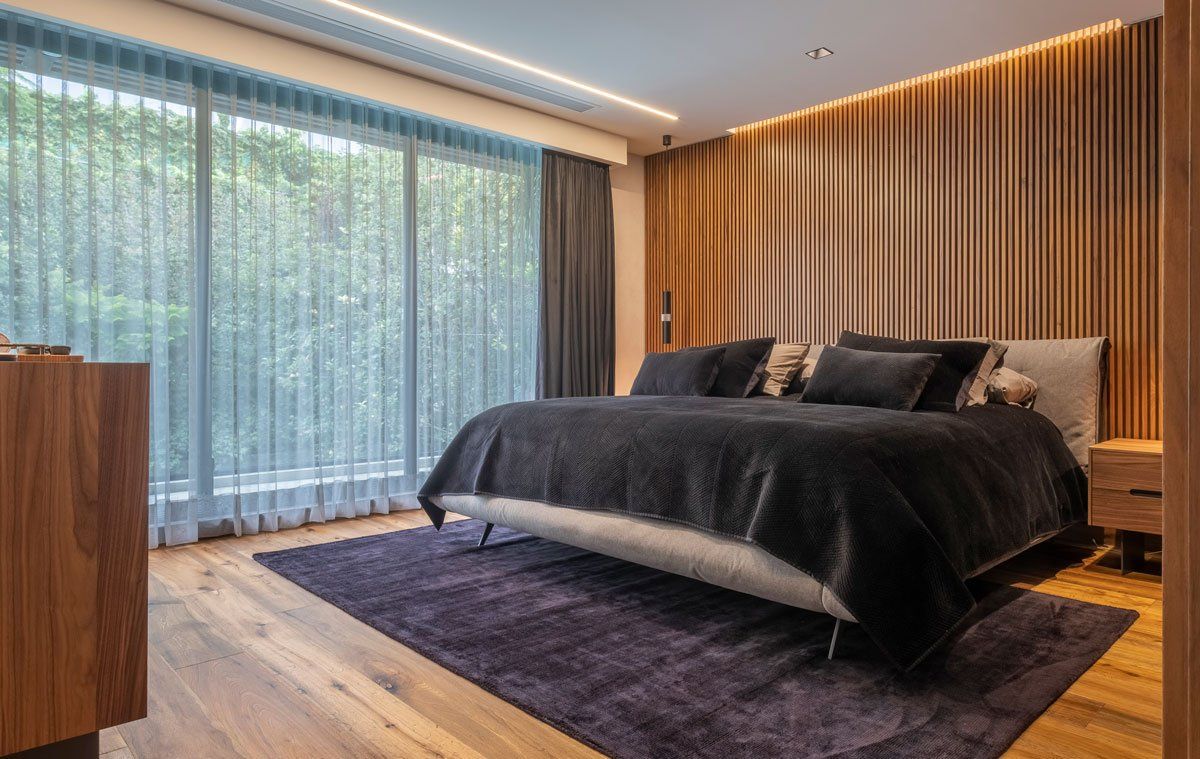 Dormitorio moderno con cama oscura, alfombra morada, suelo de madera y ventanas de suelo a techo con cortinas.
