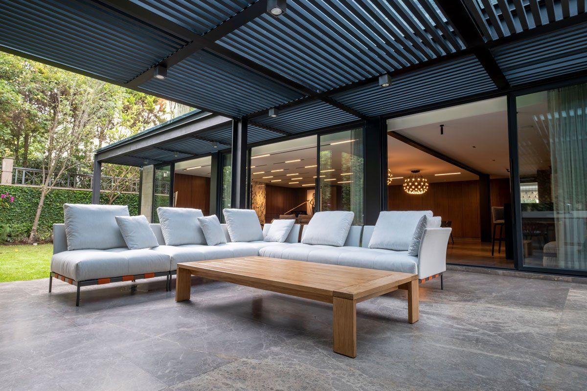 Patio exterior con sillones grises, mesa de centro de madera y casa moderna con paredes de cristal.