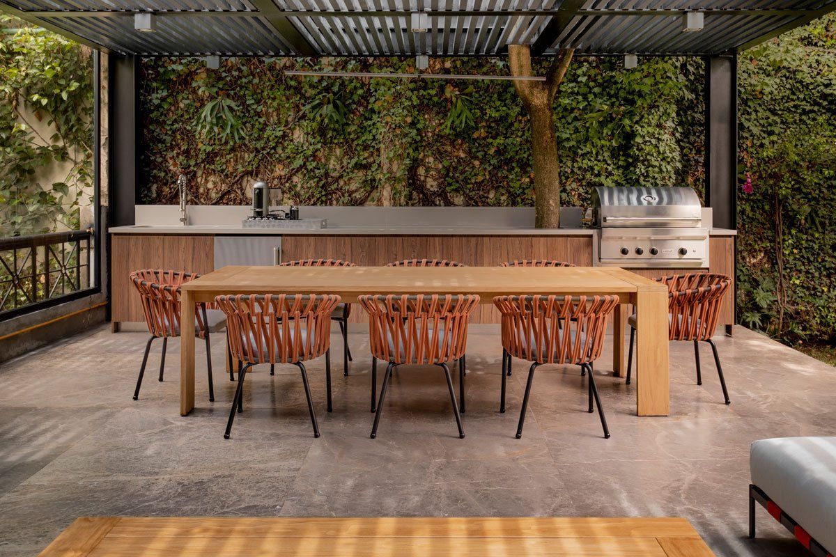 Cocina y comedor al aire libre con mesa larga de madera, taburetes y barra bajo una pérgola.