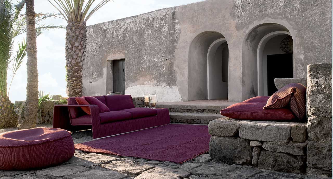 Patio exterior con asientos morados, una mesa de piedra y paredes arqueadas de estuco blanco bañadas por la luz del sol.