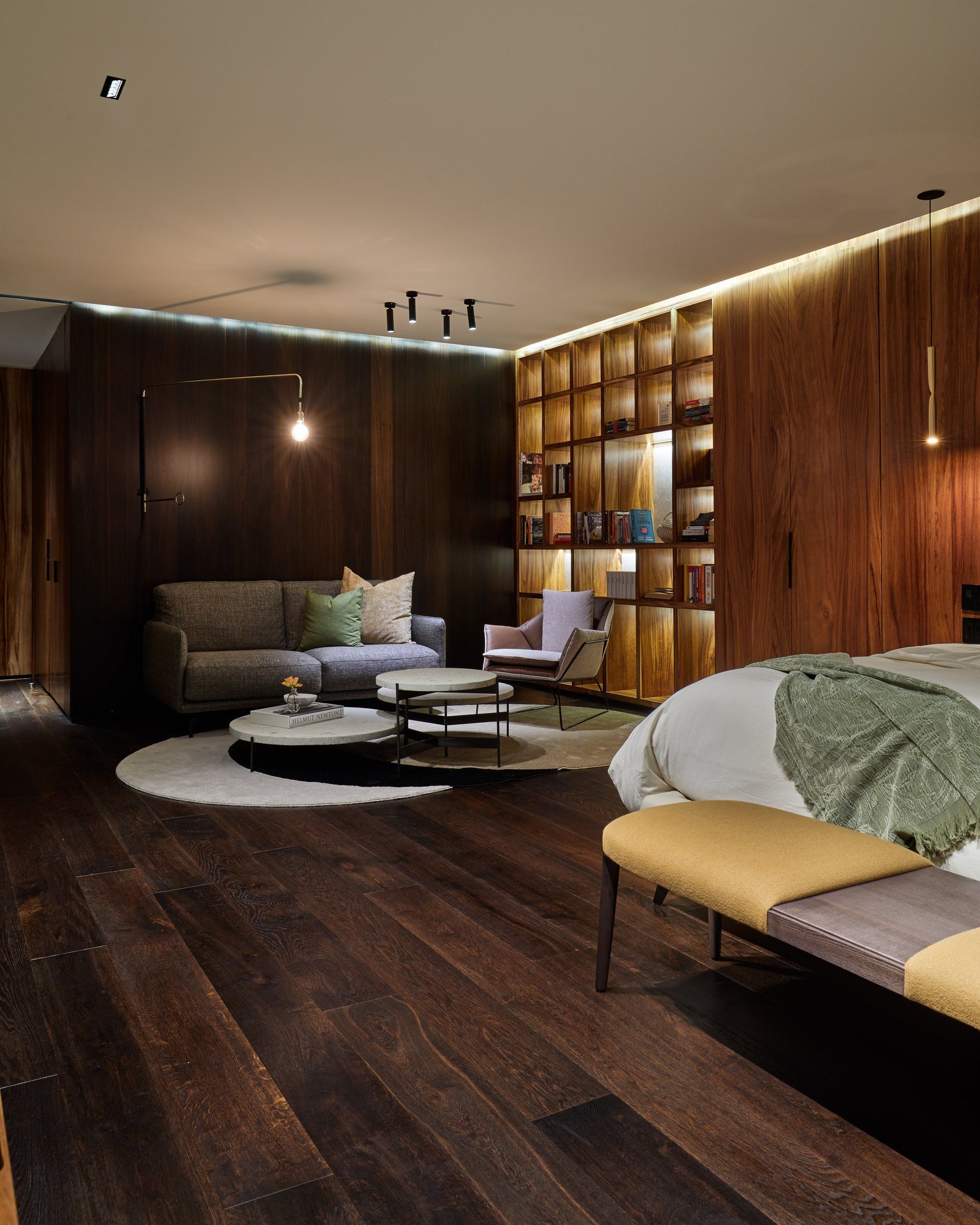 Dormitorio moderno con suelos de madera oscura, iluminación cálida, sofá, estanterías y una cama con ropa de cama verde.
