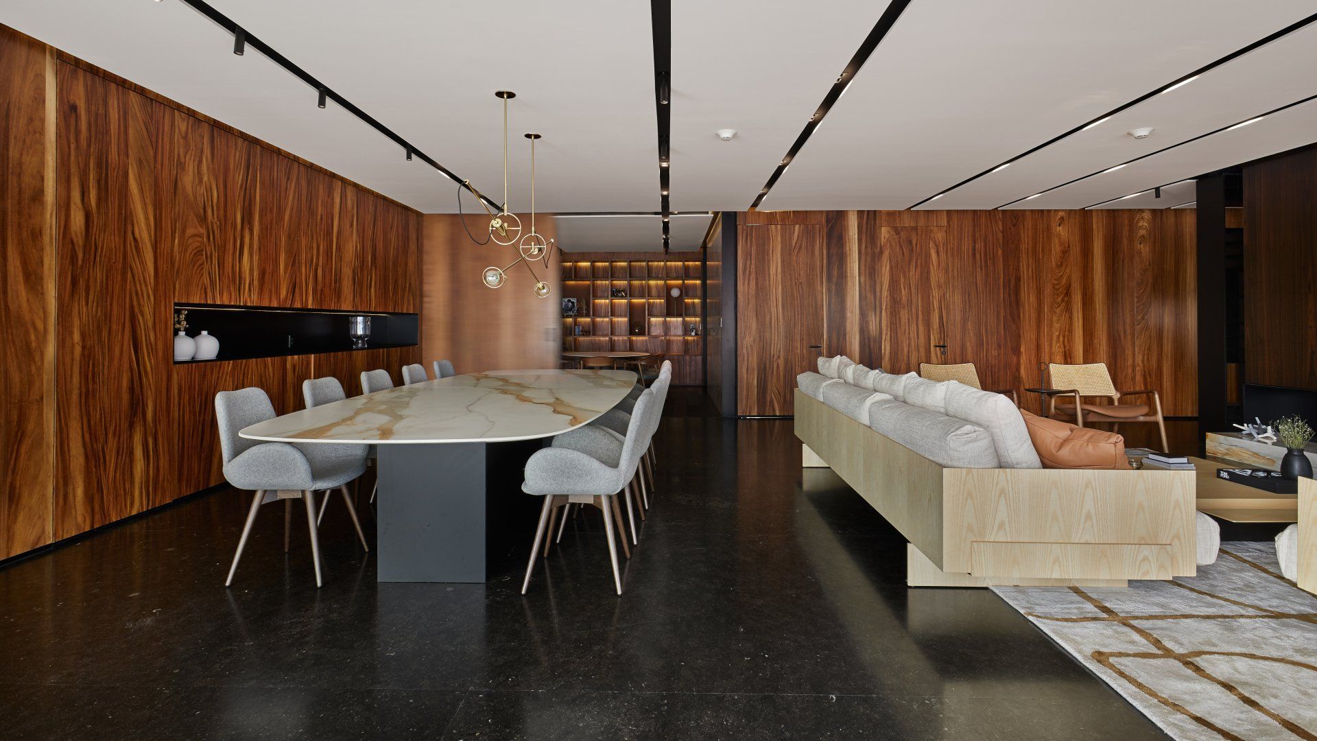 Sala de conferencias moderna con mesa larga, sillas blancas, sofás beige, paredes revestidas de madera y suelo oscuro.