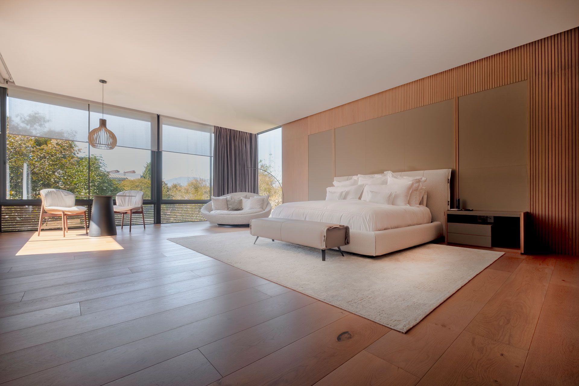 Dormitorio moderno con una cama grande, detalles en madera y ventanales que van del suelo al techo con vistas a los árboles.