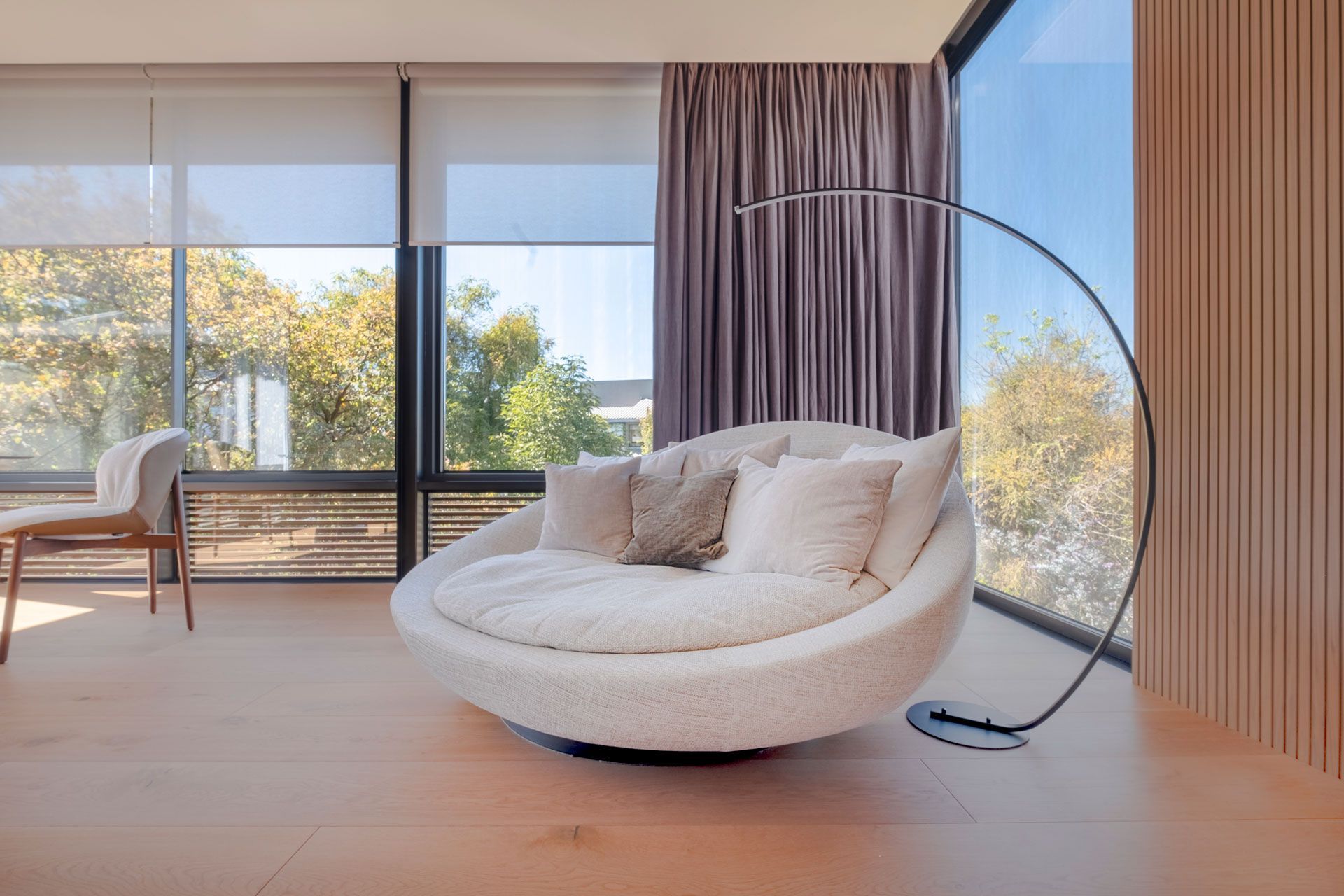 Sala de estar moderna con una silla blanca, lámpara de pie y grandes ventanales con vistas a los árboles.