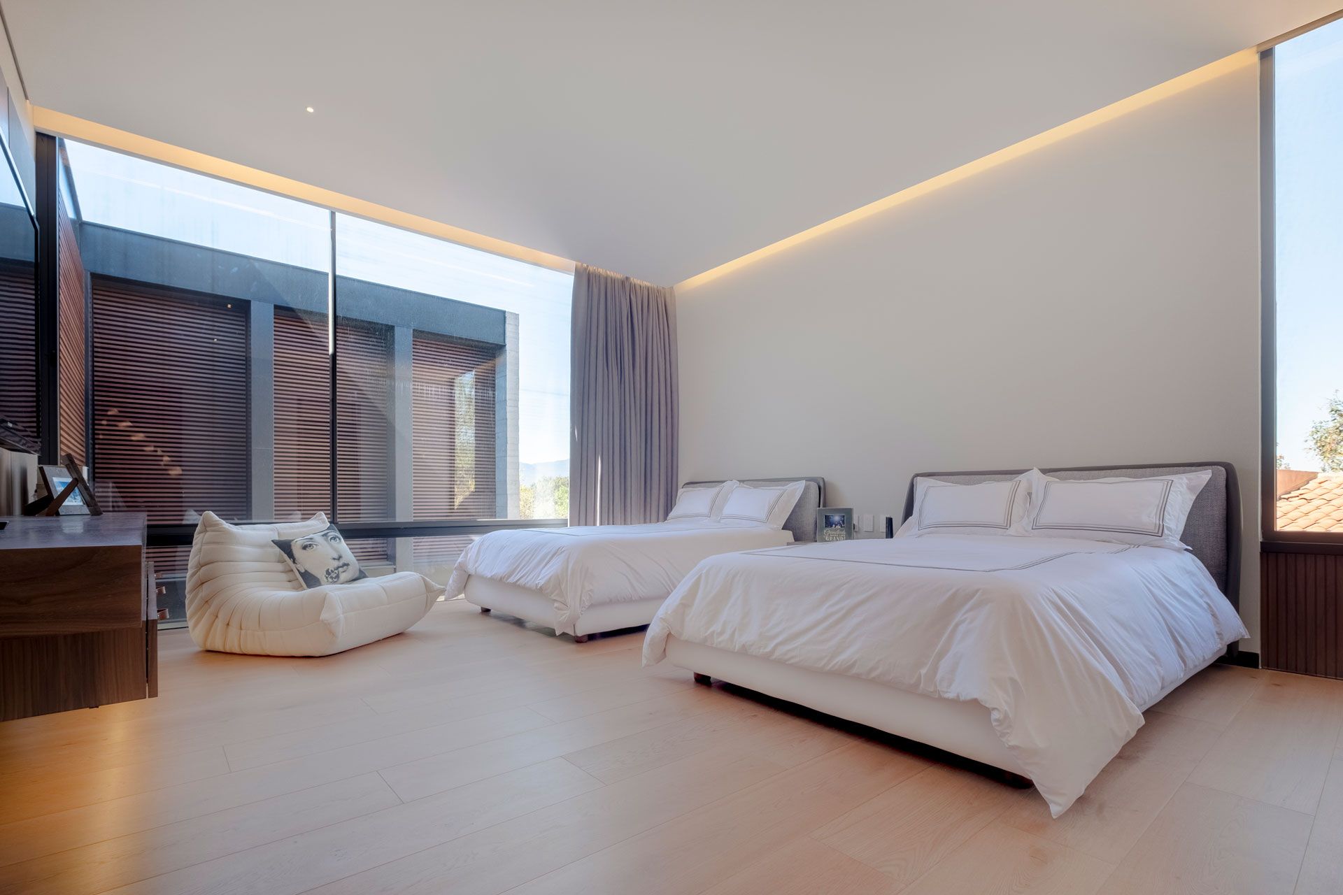 Dormitorio moderno con dos camas blancas, ventanales del suelo al techo y un acogedor rincón de lectura.