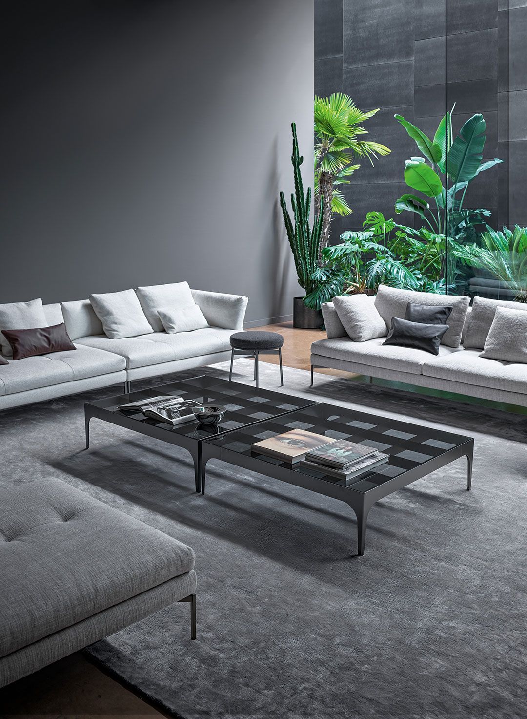 Sala de estar moderna en tonos grises con sofás, mesa de centro de cristal y plantas verdes altas cerca de una gran ventana.