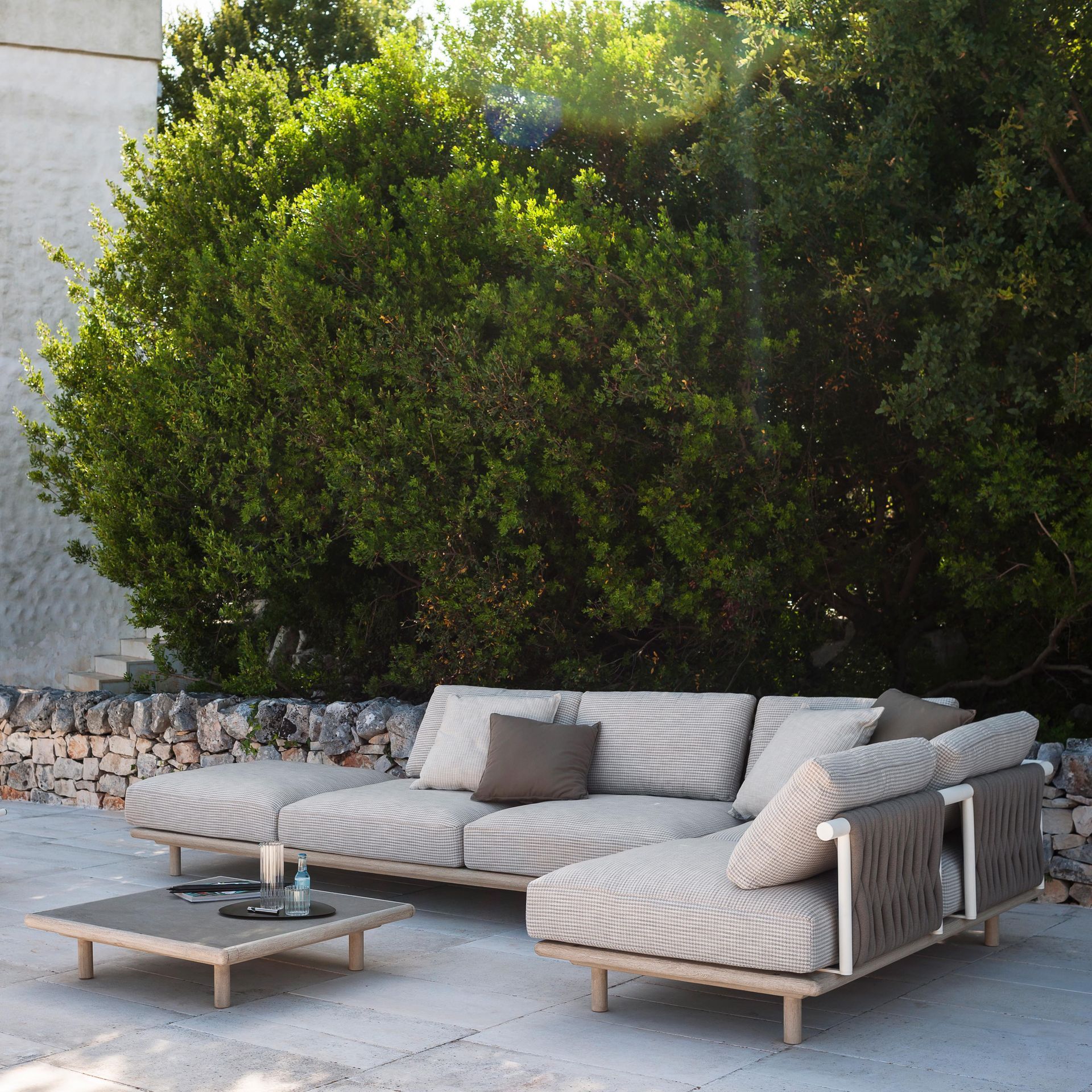 Patio exterior con un sofá seccional bajo de color beige, una mesa de centro y frondosos arbustos verdes detrás.
