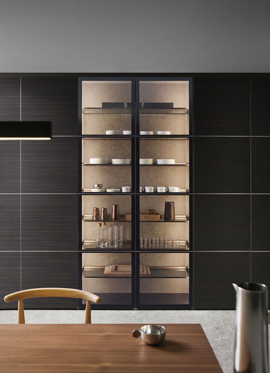 Cocina moderna con armarios oscuros, puertas de despensa de cristal y una mesa de madera en primer plano.