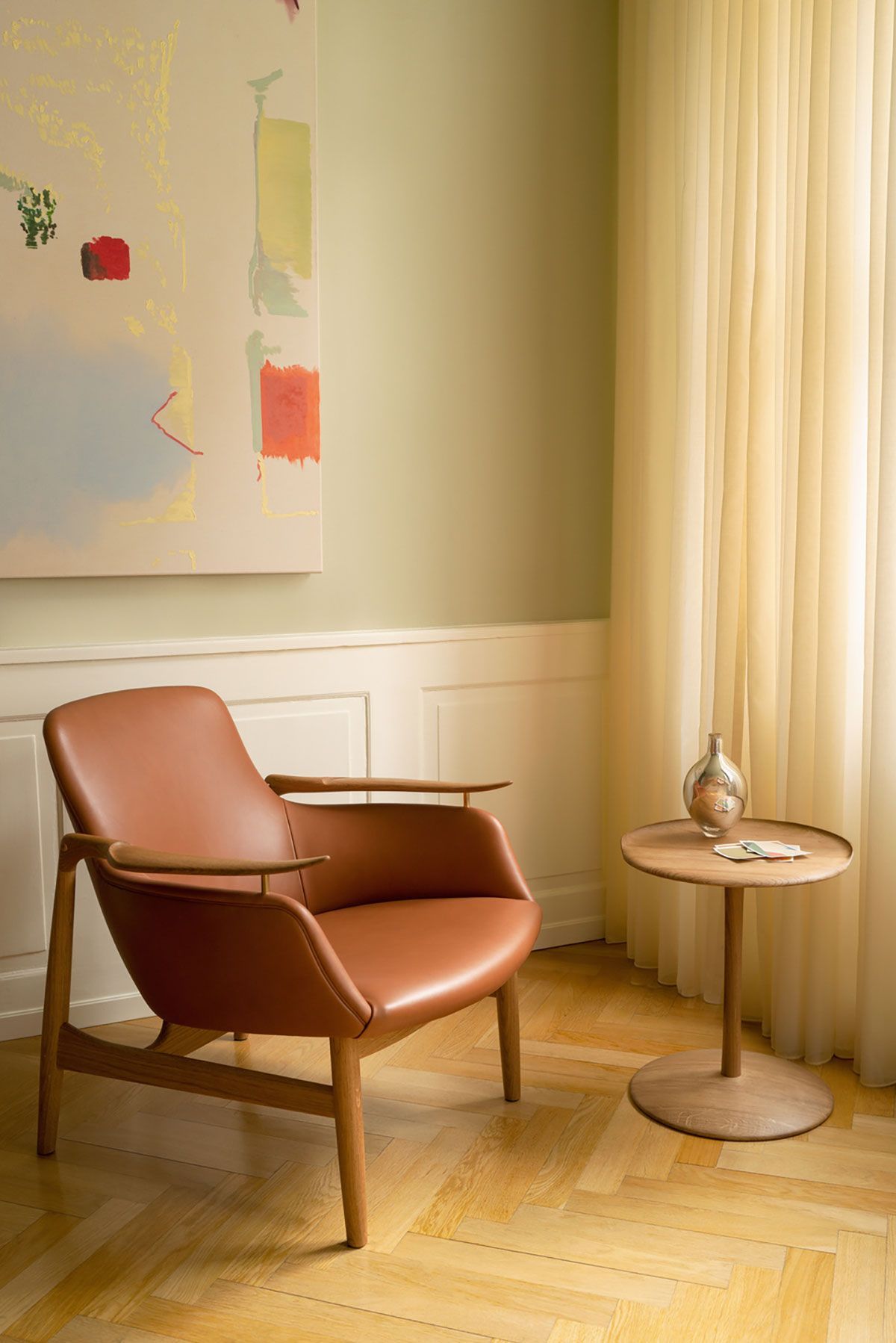 Elegante salón con sillón de cuero color camel, mesa redonda pequeña, arte abstracto y cortinas transparentes