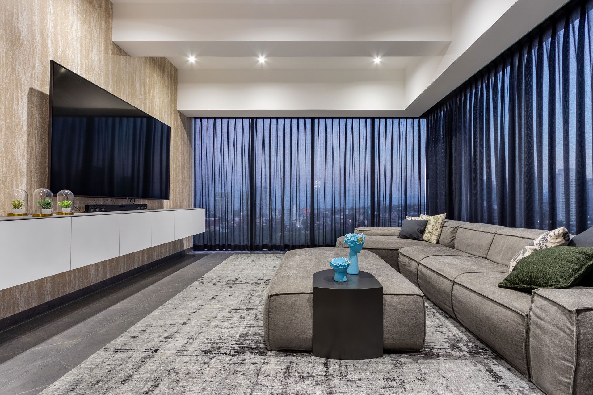 Sala de estar moderna con sofá modular gris, pared para TV, cortinas azules y alfombra estampada.