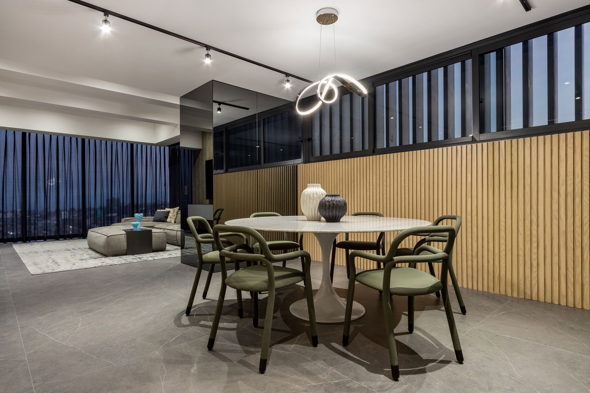 Salón moderno de planta abierta con mesa de comedor redonda, sillas verdes, sofá gris y cortinas azules.