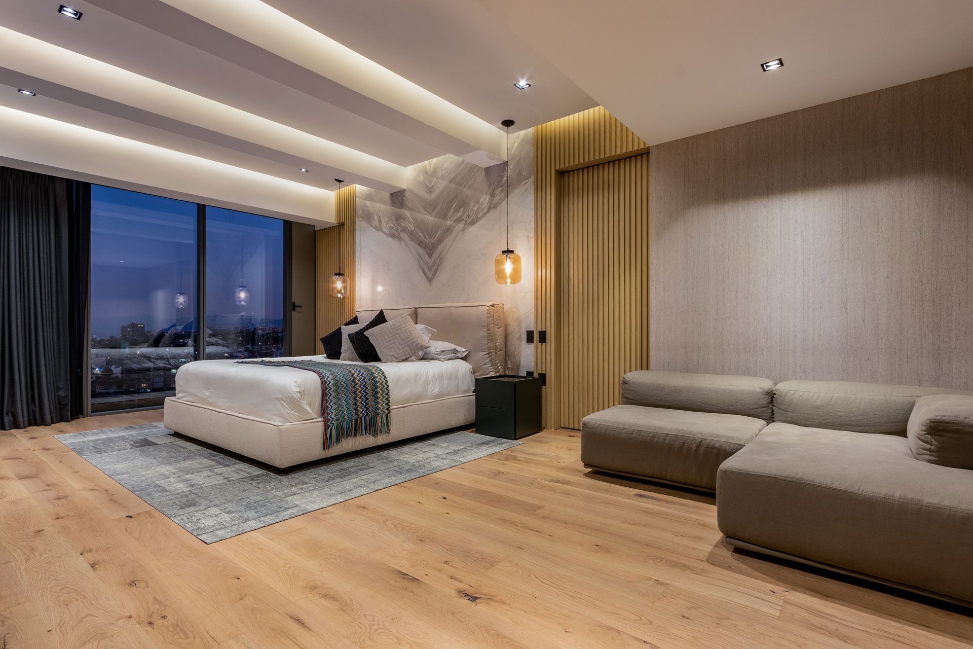 Habitación moderna con una cama grande, sofá gris, suelos de madera y vistas a la ciudad al atardecer.