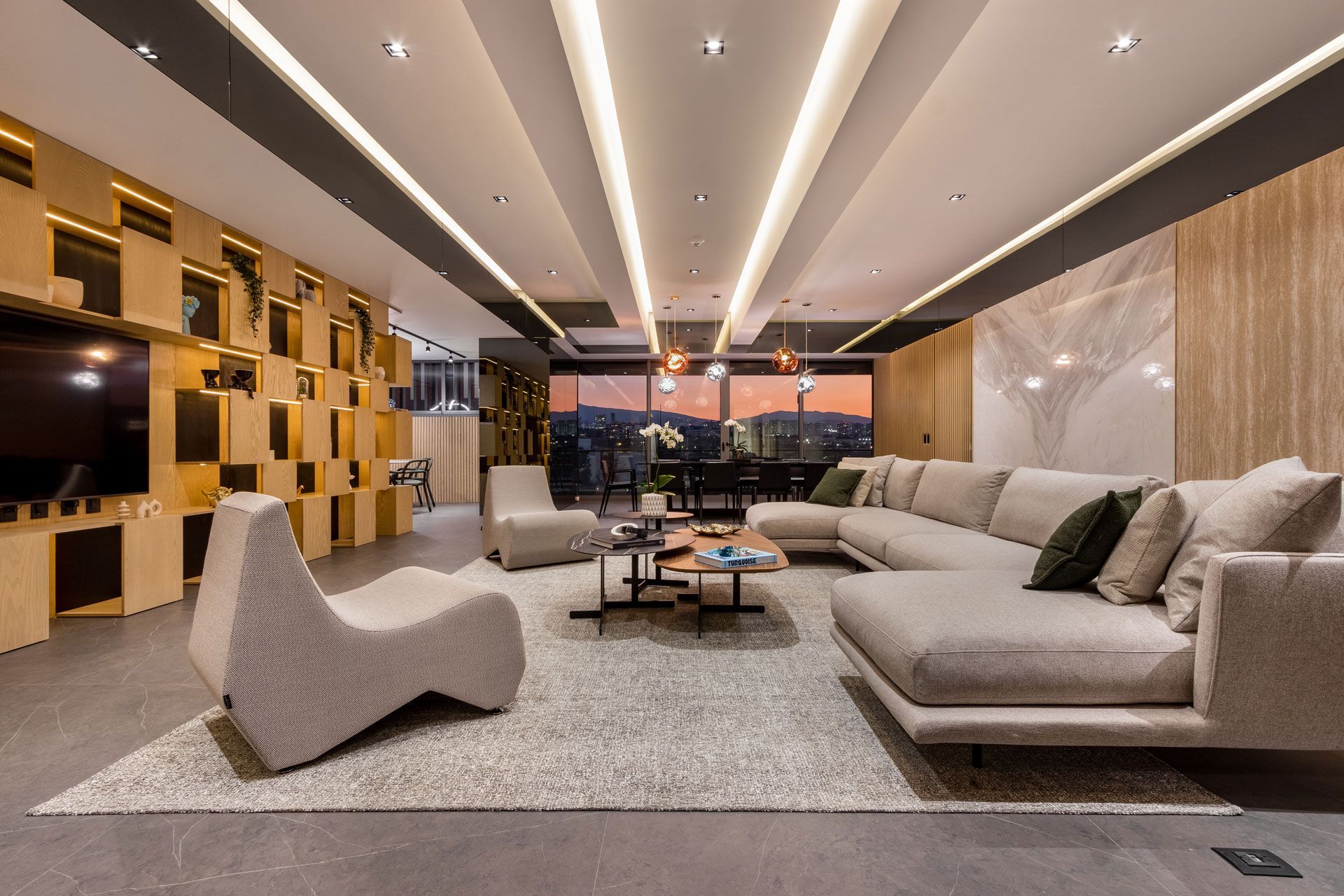 Sala de estar moderna con sofá modular beige, sillas esculturales, estanterías de madera y luces de techo cálidas.
