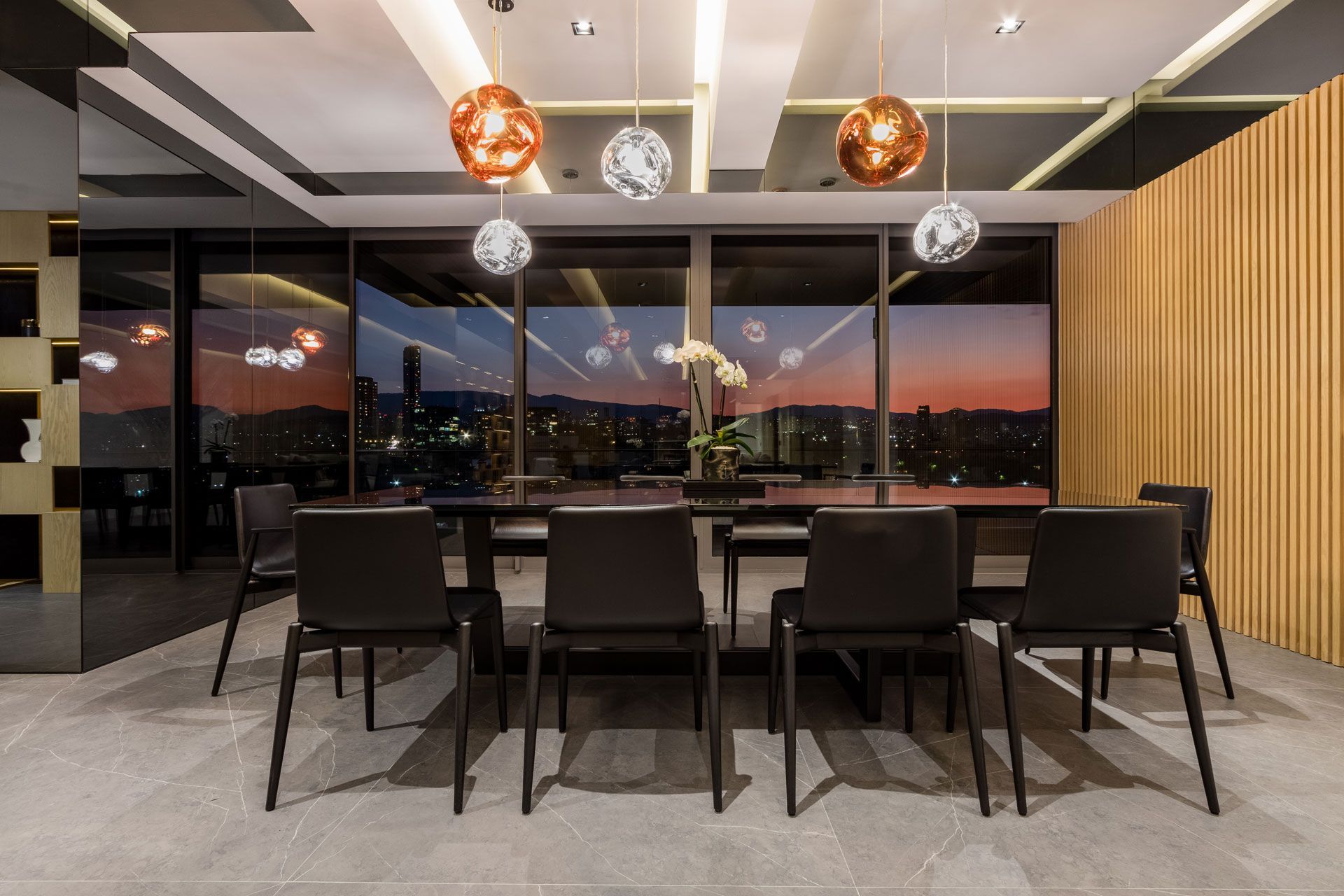 Comedor moderno con sillas negras, una mesa larga, lámparas colgantes y vistas a la ciudad al atardecer.