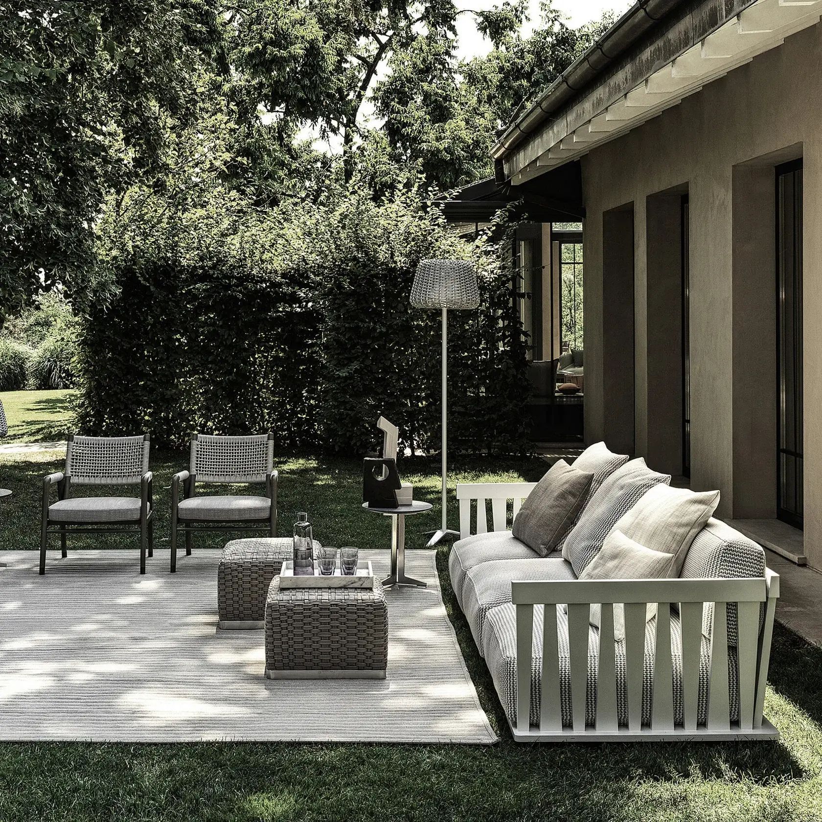 Patio con sofá blanco, dos sillas y una mesa de mimbre junto a una casa y un jardín exuberante.