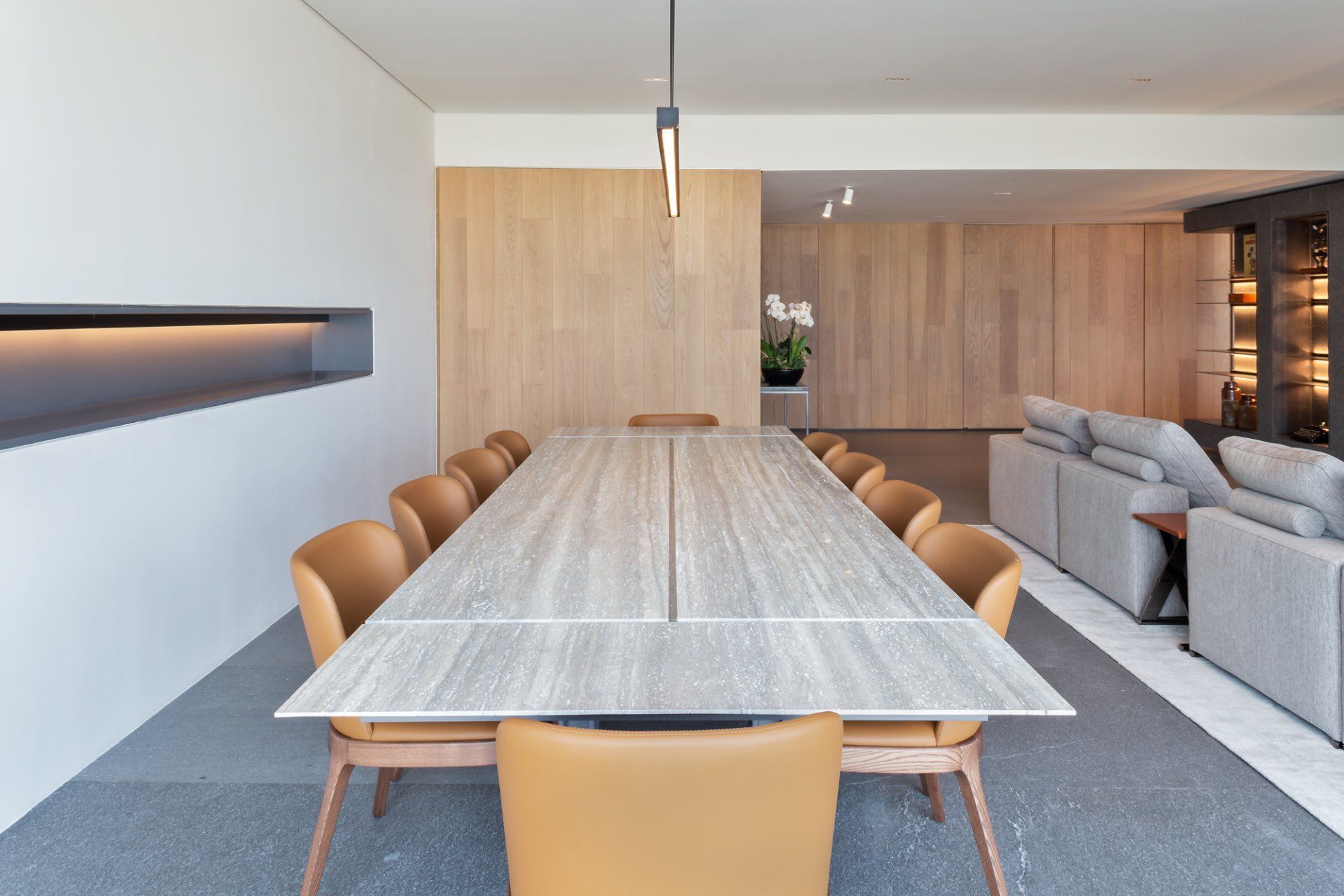 Comedor moderno con una mesa larga de madera, sillas color canela y un sofá gris claro al fondo.