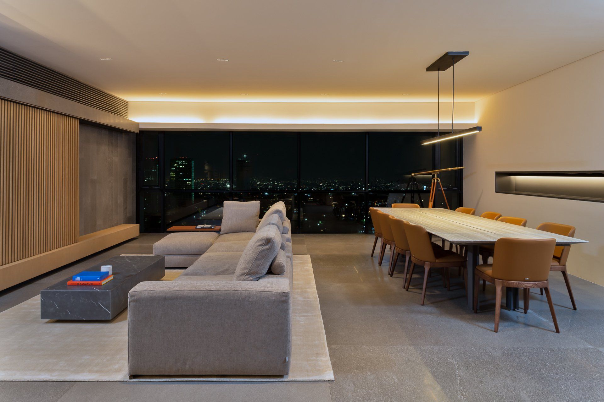 Salón-comedor moderno con sofá gris, mesa larga de madera y luces de la ciudad por la noche.