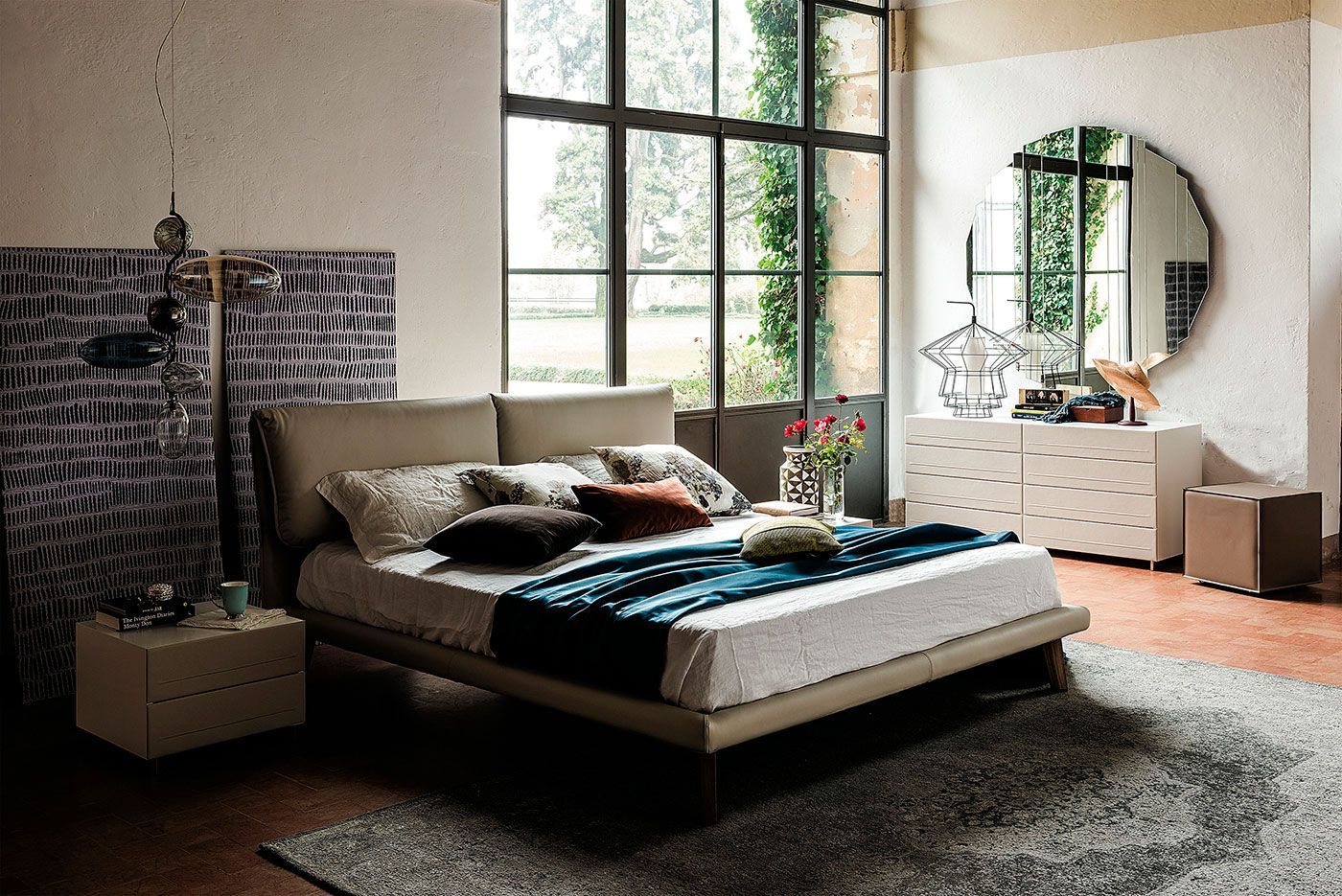Acogedor dormitorio con gran ventanal, cama, cómoda redonda con espejo y alfombra de pelo largo en tonos neutros cálidos.