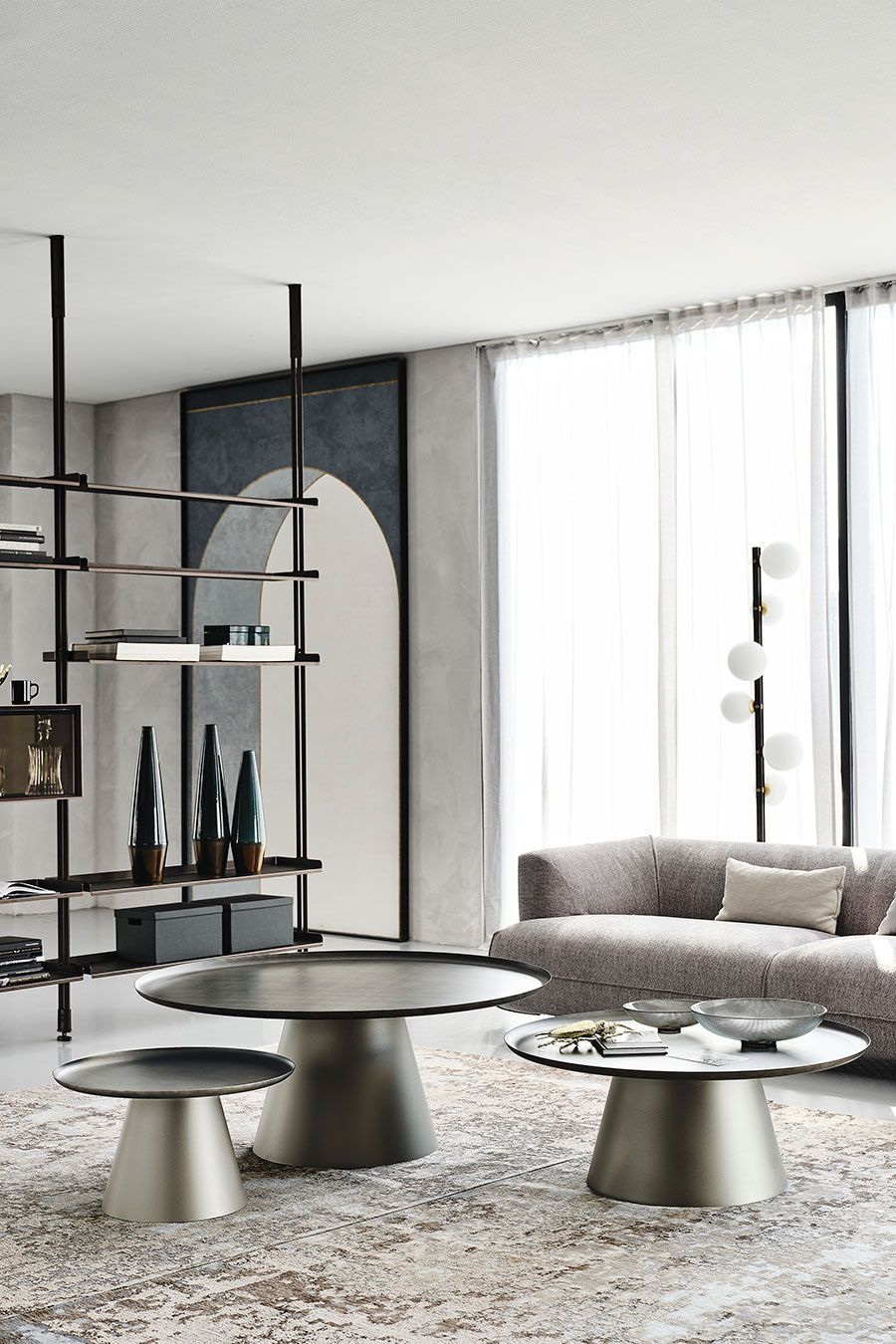 Sala de estar moderna con sofá gris, mesas de centro redondas, estanterías y grandes ventanales con cortinas transparentes.