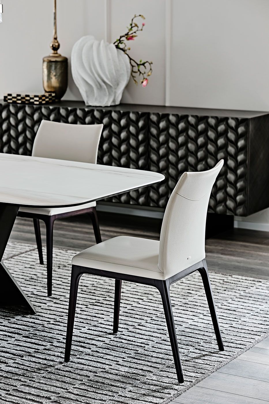 Comedor moderno con sillas negras, mesa blanca, alfombra estampada y aparador oscuro contra una pared clara.