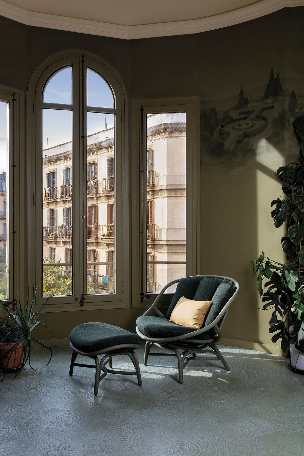 Elegante salón con altos ventanales arqueados, una silla verde, un puf y vistas a la ciudad.