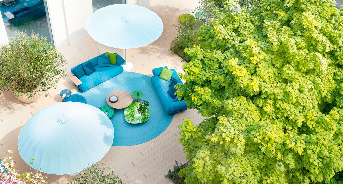 Vista aérea de una zona de estar de un patio azul bajo sombrillas blancas junto a un gran árbol verde.