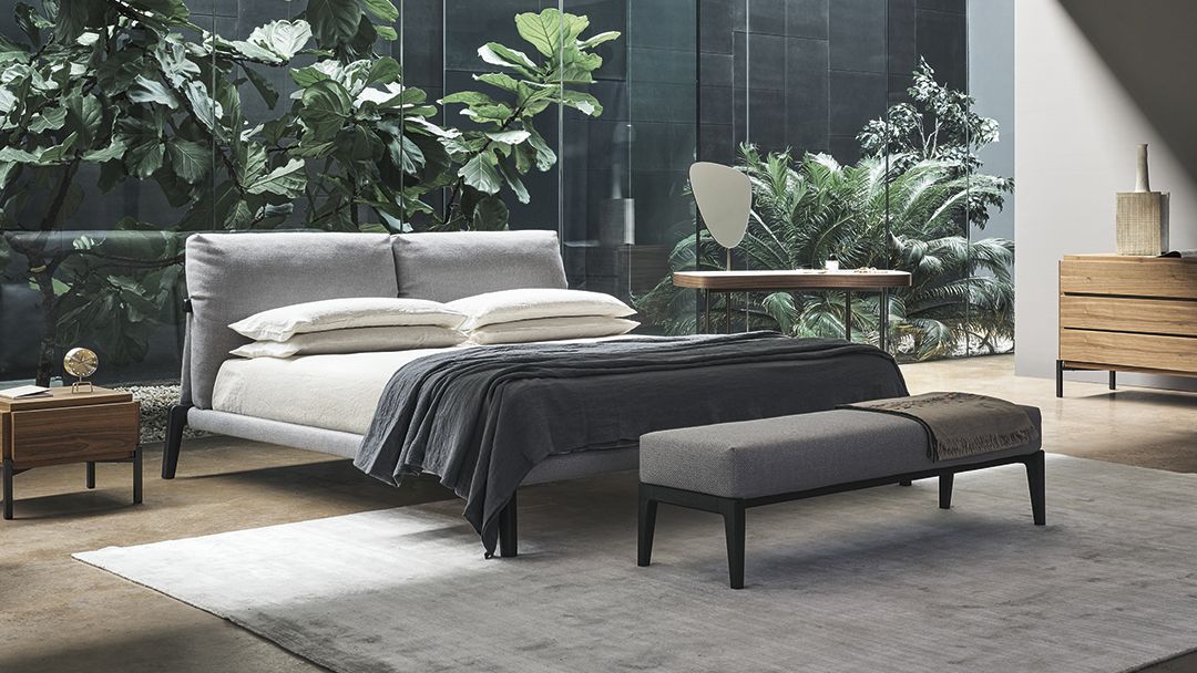 Dormitorio moderno con cama baja, banco y ventanales que van del suelo al techo con vistas a la vegetación.