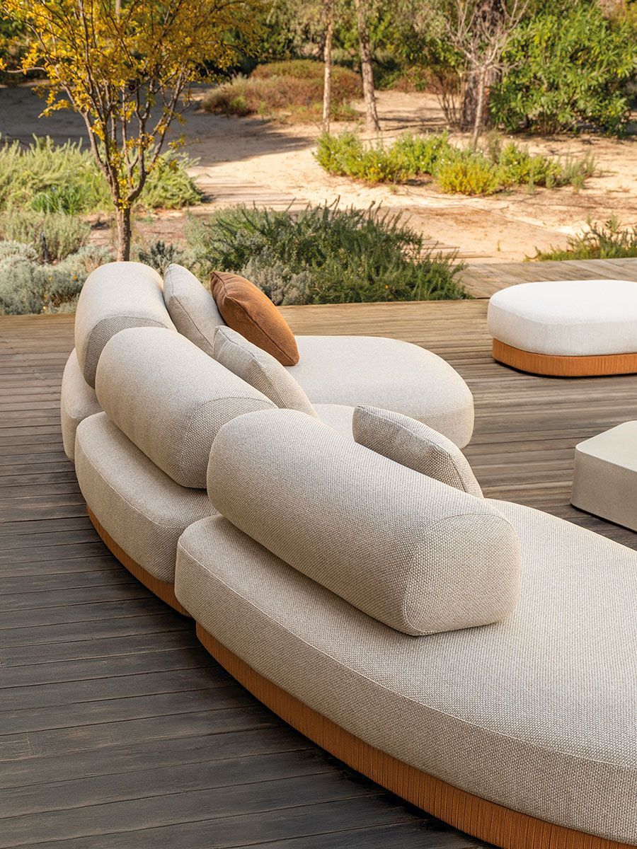 Tumbona curva de color beige para exteriores, situada en una terraza de madera junto a un sendero ajardinado y vegetación.