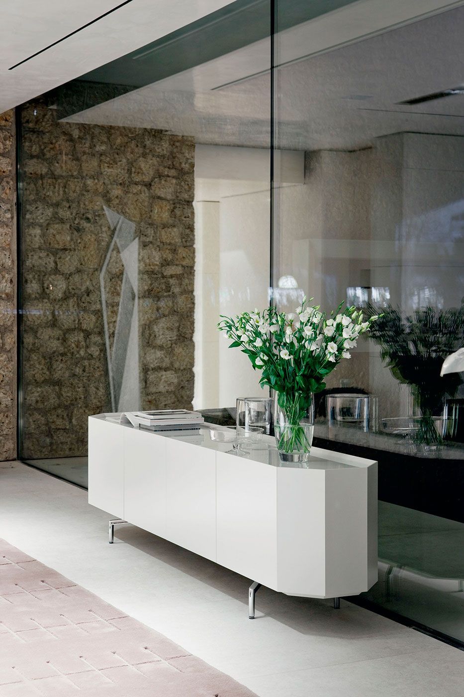 Vestíbulo de oficina moderno con consola blanca, pared de vidrio, pared decorativa de piedra y un jarrón de flores blancas.
