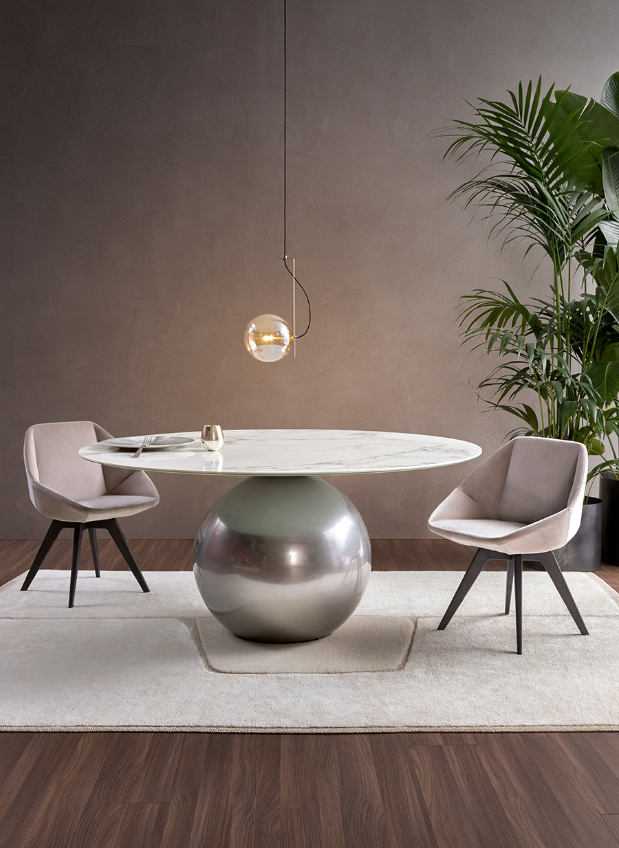 Comedor moderno con mesa redonda blanca, base de pedestal plateada, dos sillas grises y lámpara colgante.