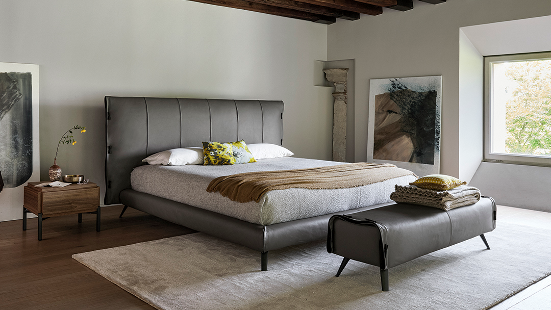 Dormitorio moderno con cama tapizada en gris, banco, mesitas de noche y grandes ventanales.