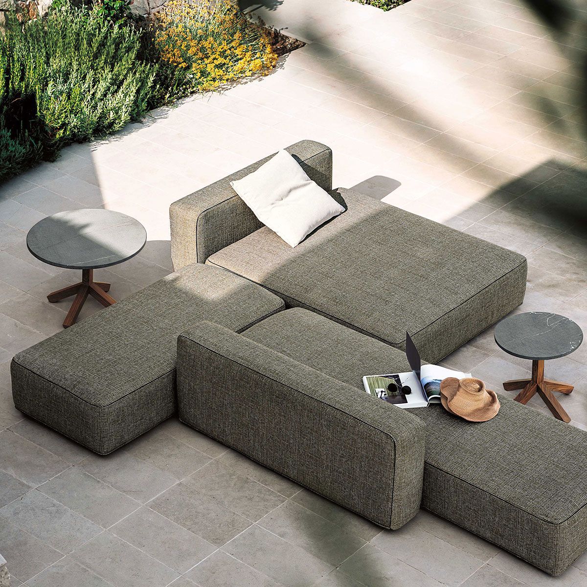 Conjunto modular de muebles de exterior grises con mesas auxiliares en un patio embaldosado, visto desde arriba.