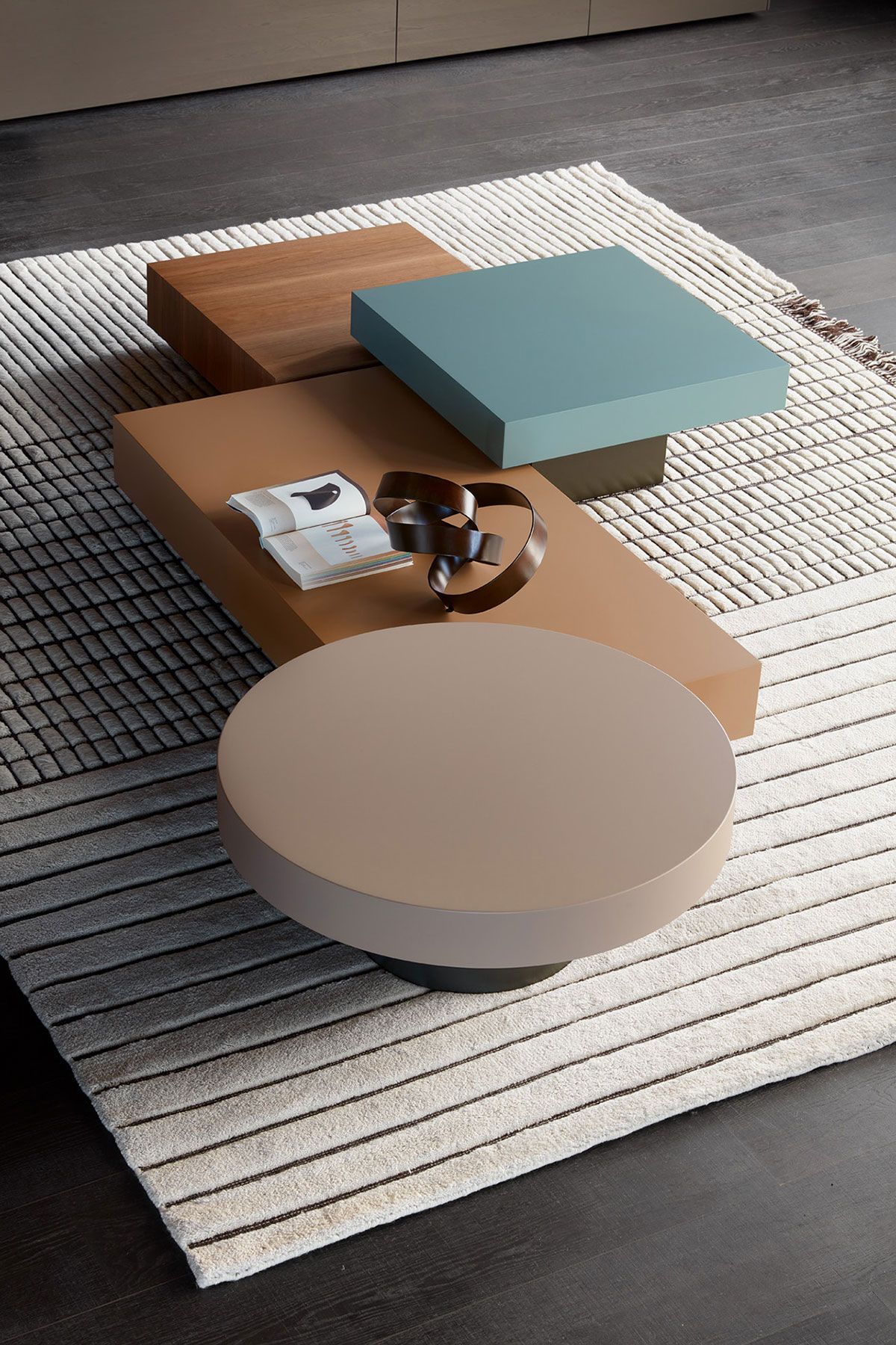 Sobre una alfombra, una mesa de centro minimalista exhibe objetos geométricos en tonos beige, verde azulado y blanco