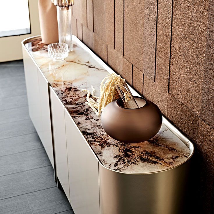 Mesa consola moderna con tablero de mármol, jarrón de bronce y decoración de cristal junto a una pared marrón texturizada.