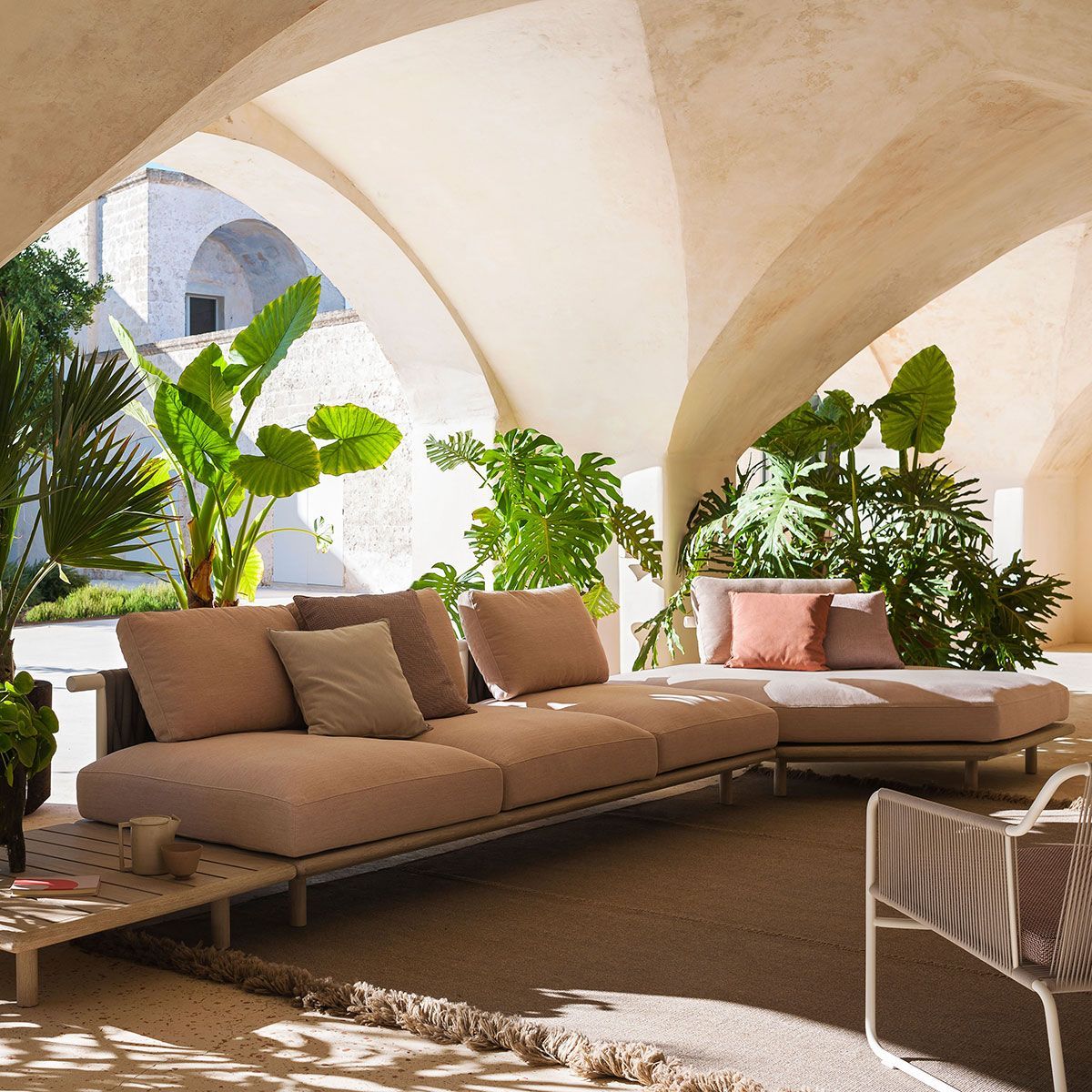 Patio soleado con sofá modular beige, arcos blancos y exuberantes plantas verdes.