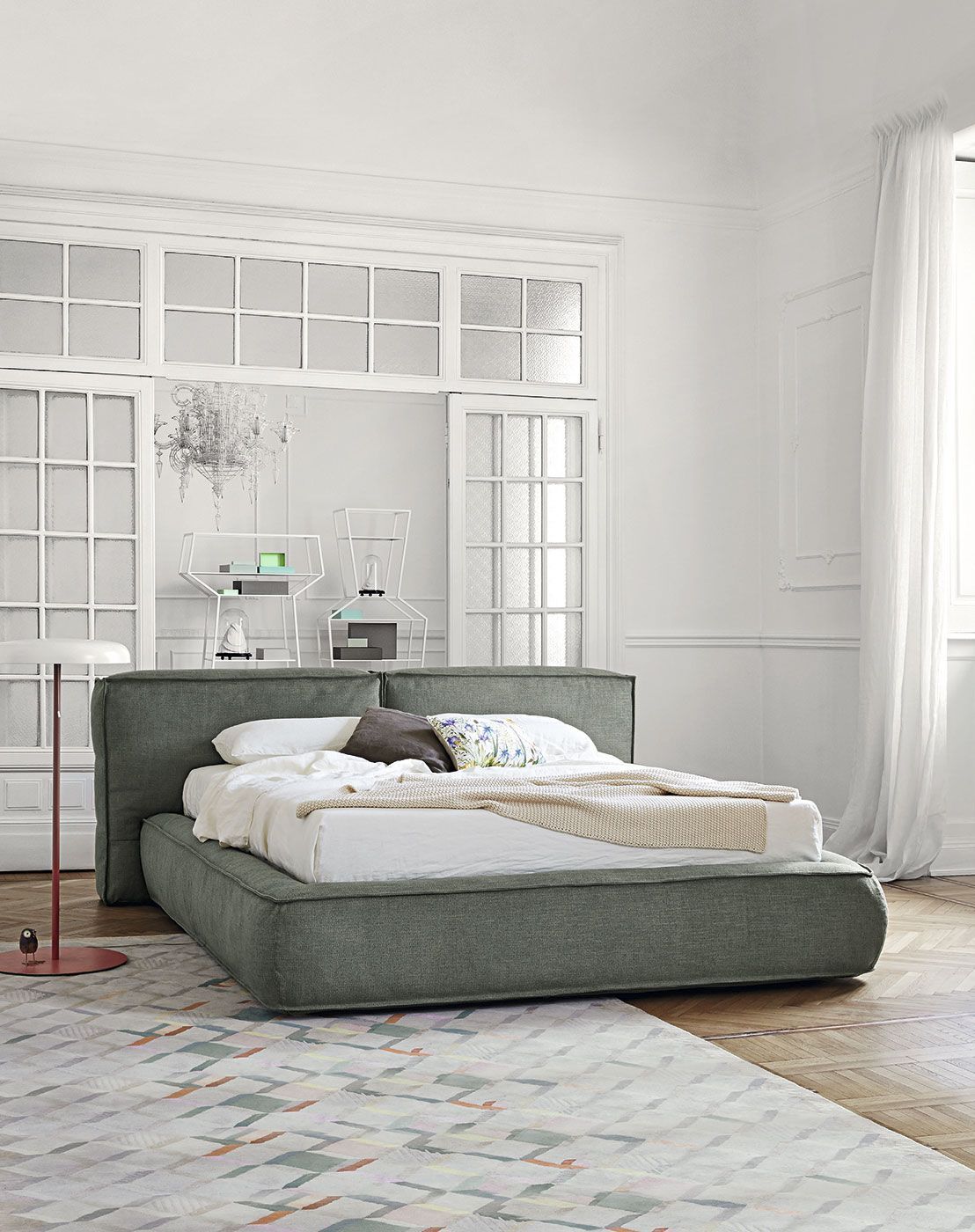 Cama tapizada en verde en un dormitorio blanco luminoso con alfombra estampada y ventanas altas.