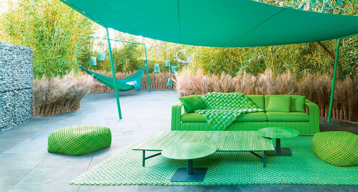 Salón exterior verde con sofá, mesa baja y toldos en un entorno de jardín soleado.