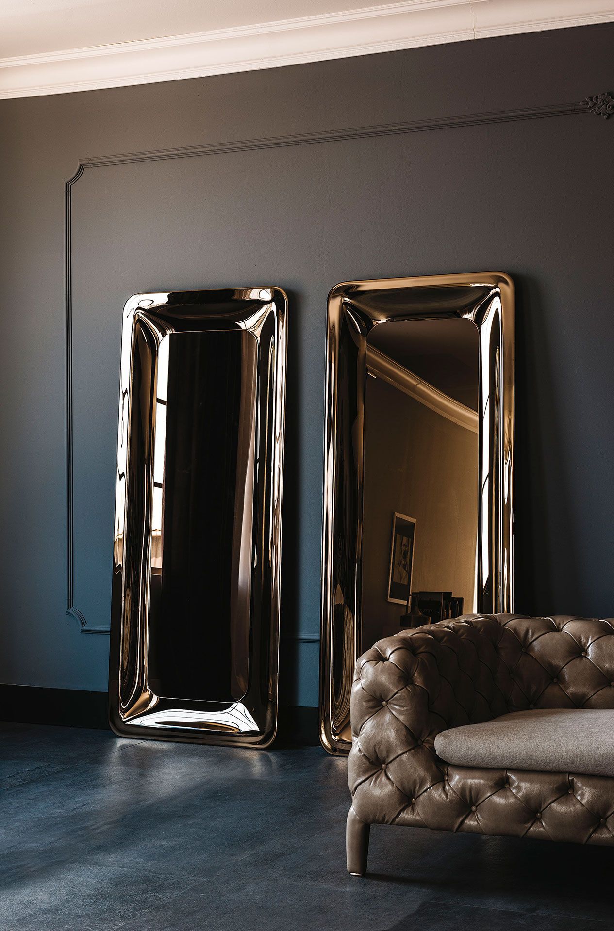 Elegante habitación con dos espejos altos con marcos dorados y un sofá de cuero capitoné contra una pared gris oscuro.