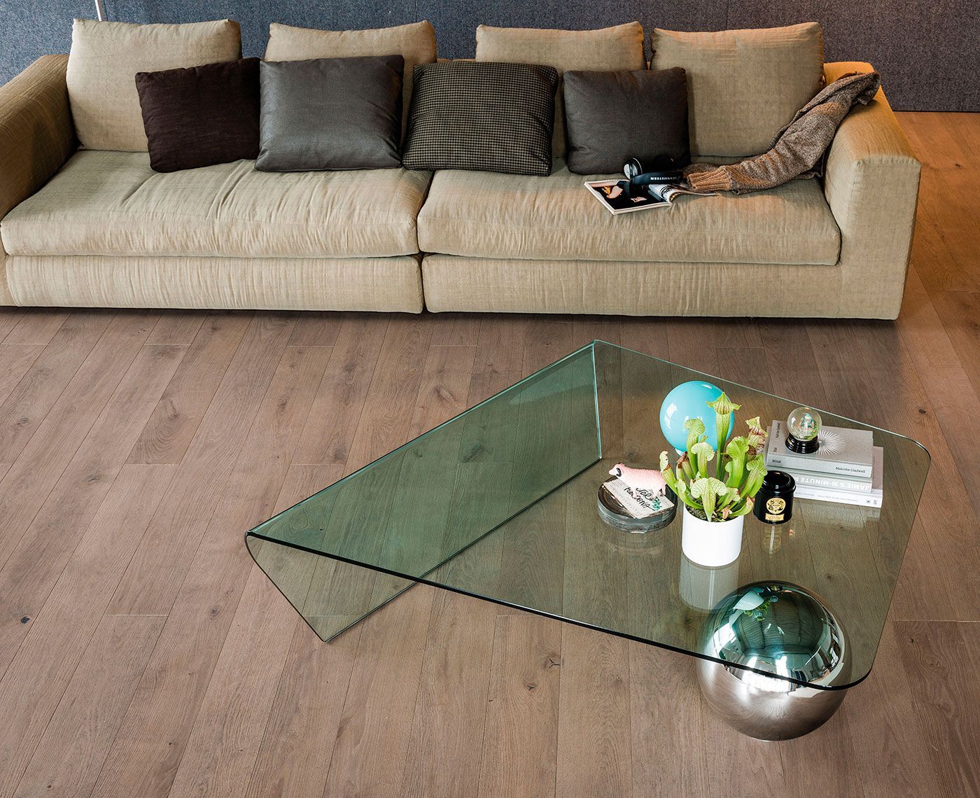 Sala de estar moderna con sofá beige, cojines oscuros y una mesa de centro de cristal sobre un suelo de madera.