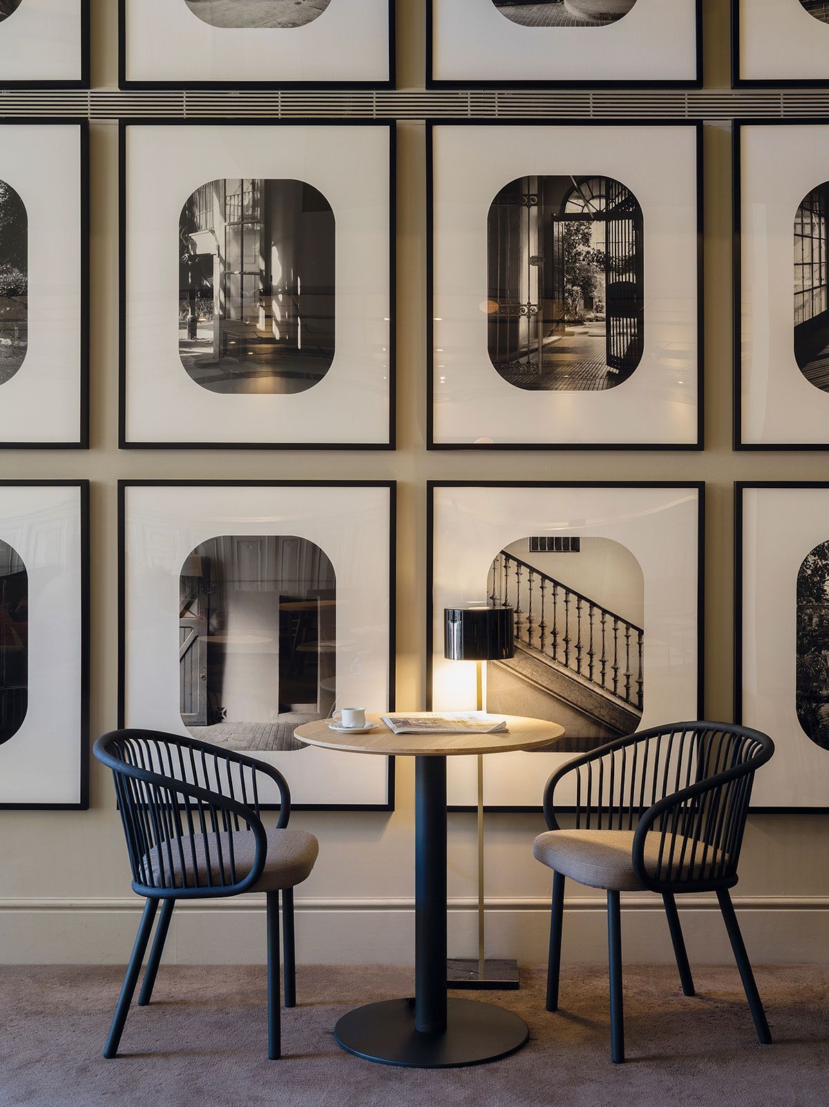 Cafetería moderna con dos sillas negras y una pequeña mesa redonda frente a una pared de fotos enmarcadas.