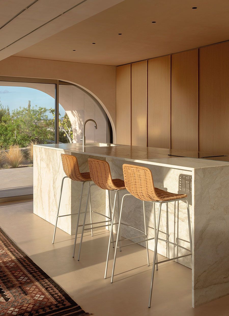 Cocina moderna con isla de mármol, tres taburetes de mimbre y ventana arqueada con vistas al desierto.