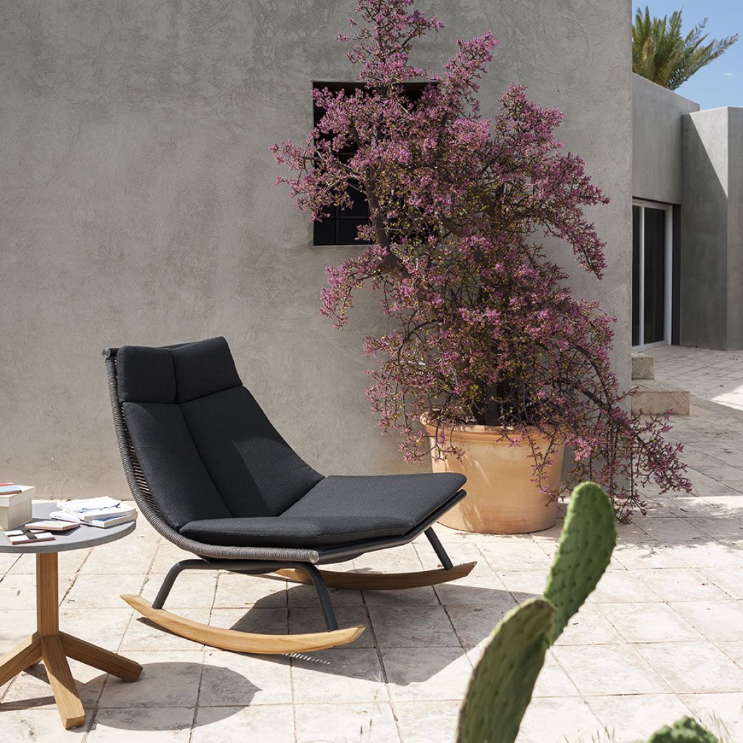 Patio moderno con una tumbona negra, una mesa pequeña y un arbusto de flores rosas junto a una pared de estuco.