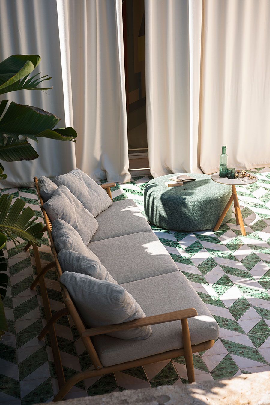 Patio soleado con un sofá beige, un puf verde, una alfombra estampada y cortinas altas junto a plantas frondosas.