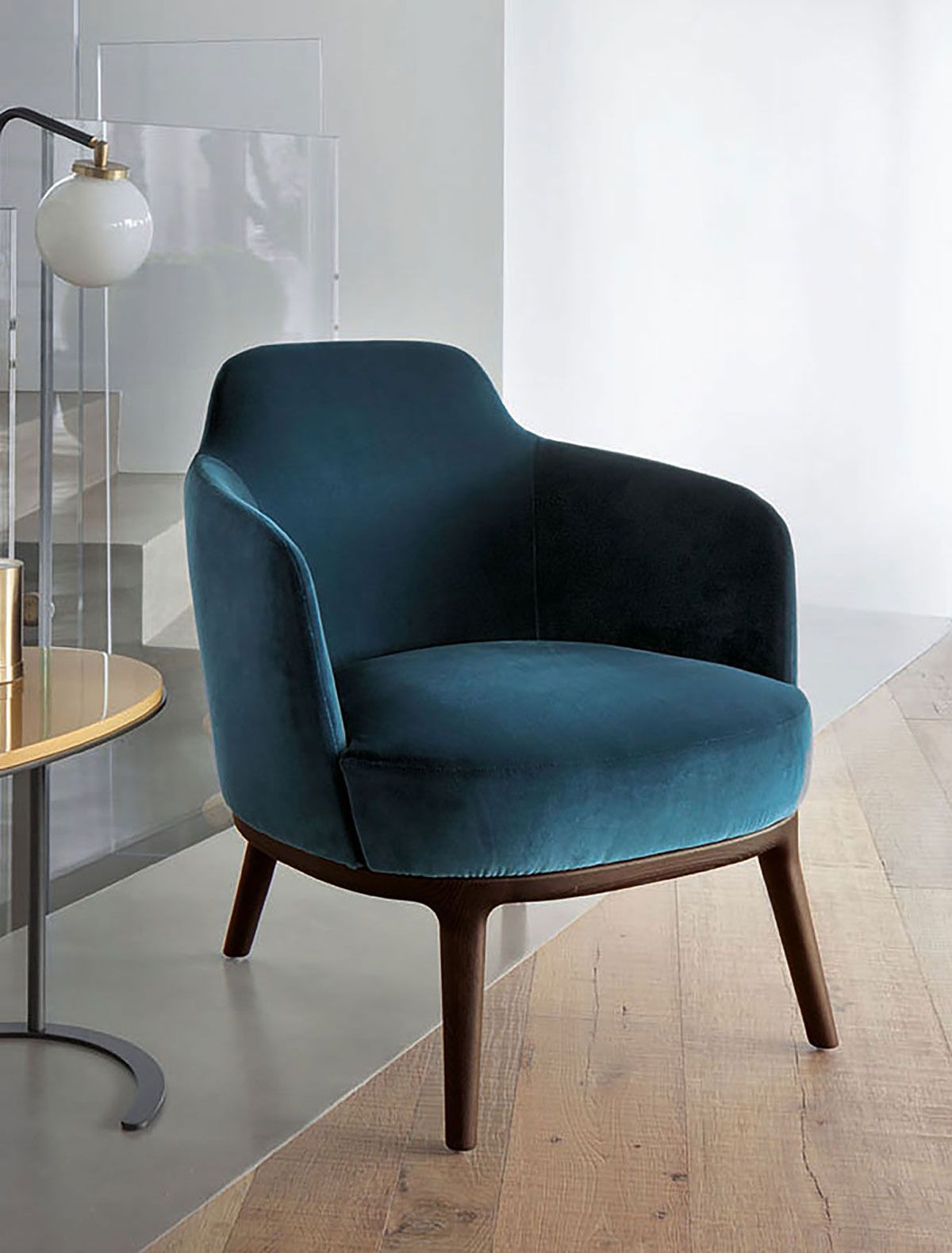 Sillón tapizado en color verde azulado con patas de madera junto a una lámpara de pie sobre un suelo de madera clara.