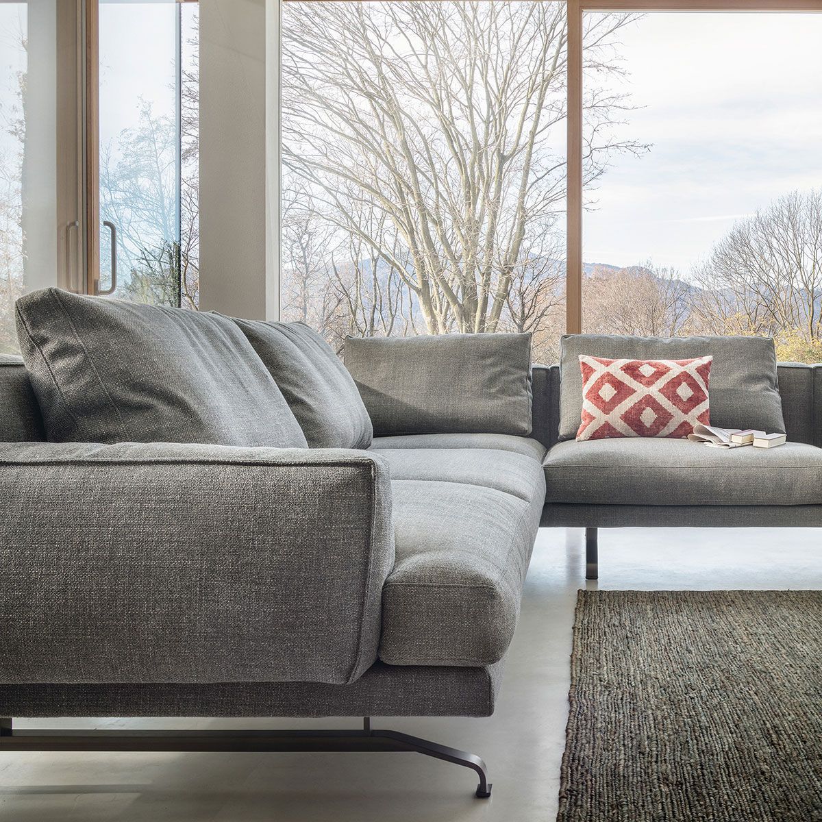 Sofá modular moderno de color gris junto a grandes ventanales, con un cojín estampado rojo y una alfombra de pelo largo