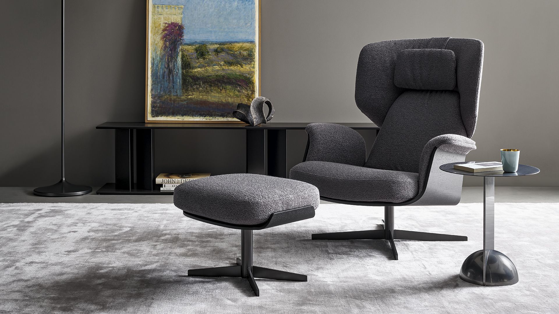 Salón moderno con sillón gris, otomana a juego, mesa auxiliar y obra de arte en una pared oscura.